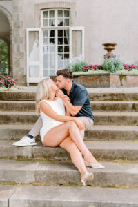 Glen Manor House Portsmouth RI | Deirdre Photography Timeless and Organic Moments | A couple sits affectionately on stone steps outside a building with open French doors. The setting is romantic, with flowers and a gentle, loving atmosphere.