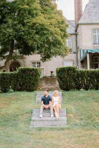 Glen Manor House Portsmouth RI | Deirdre Photography Timeless and Organic Moments | A couple sits on stone steps in front of a large, historic house with manicured hedges. The greenery and cozy setting convey a serene, romantic atmosphere.