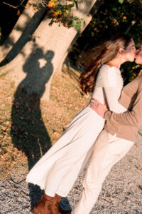 Blithewold Mansion, Gardens and Arboretum | Deirdre Photography Timeless and Organic Moments | A couple kisses under autumn trees, their shadow cast on a tree trunk. She wears a white dress, he wears beige. Leaves and sunlight create a warm, romantic scene.
