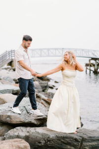 Glen Manor House Portsmouth RI | Deirdre Photography Timeless and Organic Moments | A couple stands on large rocks by the water; the woman in a white dress smiles as she holds hands with the man in a shirt and jeans. A bridge is in the background.