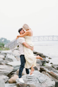 Glen Manor House Portsmouth RI | Deirdre Photography Timeless and Organic Moments | A couple embraces joyfully on rocky shore, woman in white dress lifted by man in casual wear. Background features a misty bridge over calm water. Romantic mood.