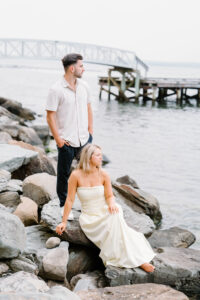 Glen Manor House Portsmouth RI | Deirdre Photography Timeless and Organic Moments | A couple poses on coastal rocks near the water, with a woman seated in a white dress and a man in a light shirt standing. A serene pier is in the background.