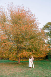 Blithewold Mansion, Gardens and Arboretum | Deirdre Photography Timeless and Organic Moments | A couple stands under a large tree with orange leaves in a grassy field. The scene is serene and autumnal, with a soft, warm ambiance.