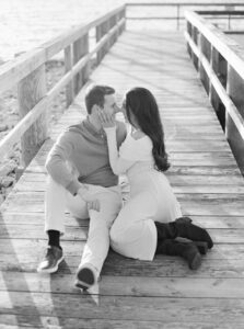 Blithewold Mansion, Gardens and Arboretum | Deirdre Photography Timeless and Organic Moments | A couple sits on a wooden pier, embracing lovingly. The woman, in a light dress, gently touches the man's face. The scene conveys romance and serenity.