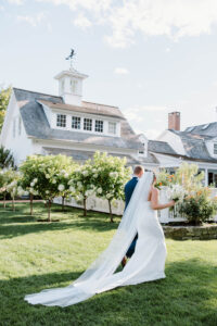 Smith Farm Garden Wedding | Deirdre Photography Timeless and Organic Moments | A bride in a long white gown and veil walks with a groom in blue suit toward a charming white house with lush green lawn and blooming flowers.