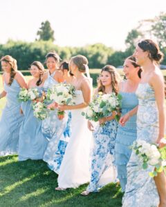 The Chanler at Cliff Walk Wedding | Deirdre Photography Timeless and Organic Moments | A bride in a white dress walks joyfully with bridesmaids in blue dresses. Each holds a bouquet of white and green flowers on a sunny day, exuding happiness.