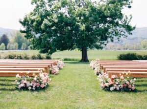 Rhode Island Wedding Photographer | Deirdre Photography Timeless and Organic Moments | Outdoor wedding setup with wooden benches adorned with pink and white flowers, set on a grassy field. A large tree stands in the background. Peaceful ambiance.