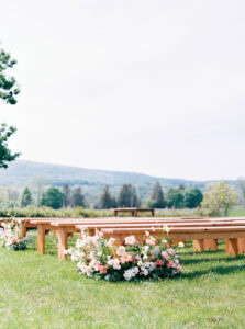 Newport RI Mansion Wedding | Deirdre Photography Timeless and Organic Moments | Outdoor wedding setup with wooden benches on a grassy field, adorned with pink and white flowers. Mountains and trees create a serene backdrop.