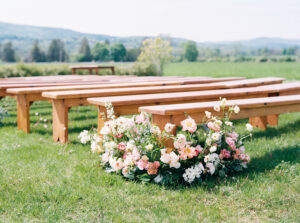 Lion Rock Farm Wedding | Deirdre Photography Timeless and Organic Moments | Wooden benches in a grassy outdoor setting adorned with pink and white flowers. A serene landscape with distant hills under a clear sky.