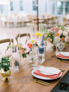 Lion Rock Farm Wedding | Deirdre Photography Timeless and Organic Moments | Elegant table setting with pink and white flowers, clear glass vases, blue candles, and coral napkins on white plates. Bright and inviting atmosphere.