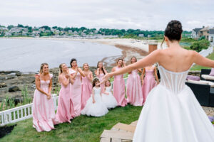 A bride opens her arms toward her bridesmaids on the lawn above the shoreline, capturing the joyful coastal energy couples love in Newport Rhode Island wedding venues.