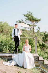 A groom in a white dinner jacket stands above the seated bride with a parasol, creating an editorial portrait on the bluff at this Newport Rhode Island wedding venue.